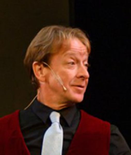 Man speaking on stage wearing a microphone headset and formal attire.