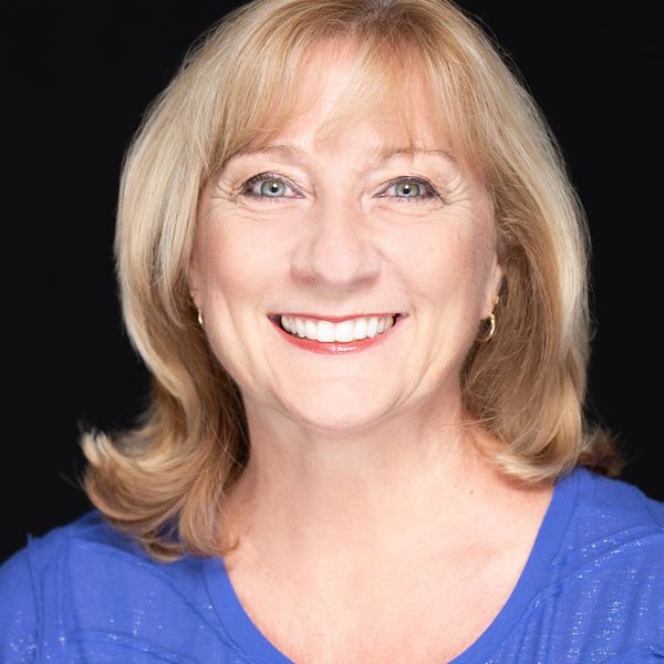 Smiling middle-aged woman with blonde hair in a blue top against a black background.