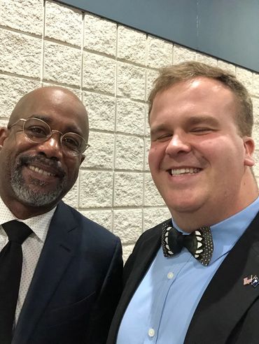Two men dressed in formal attire smiling for a selfie indoors.