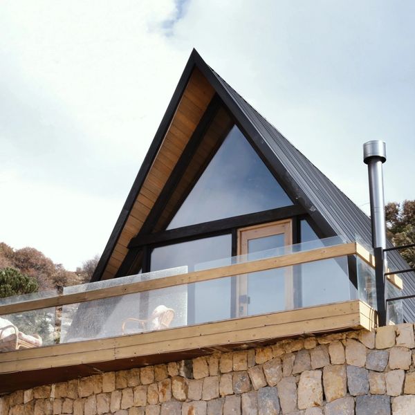 Modern A-frame cabin with glass balcony on stone foundation under blue sky.
