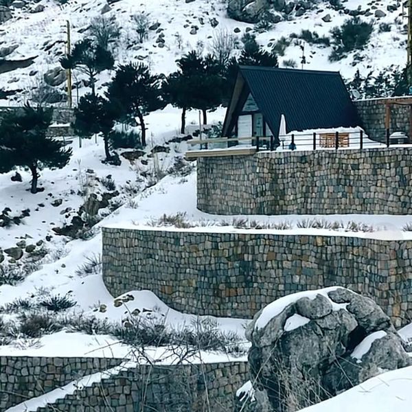 A small cabin with a black roof on a snowy hillside surrounded by stone terraces and pine trees.