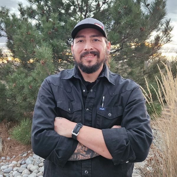 Man with beard and tattoos stands confidently outdoors with arms crossed.