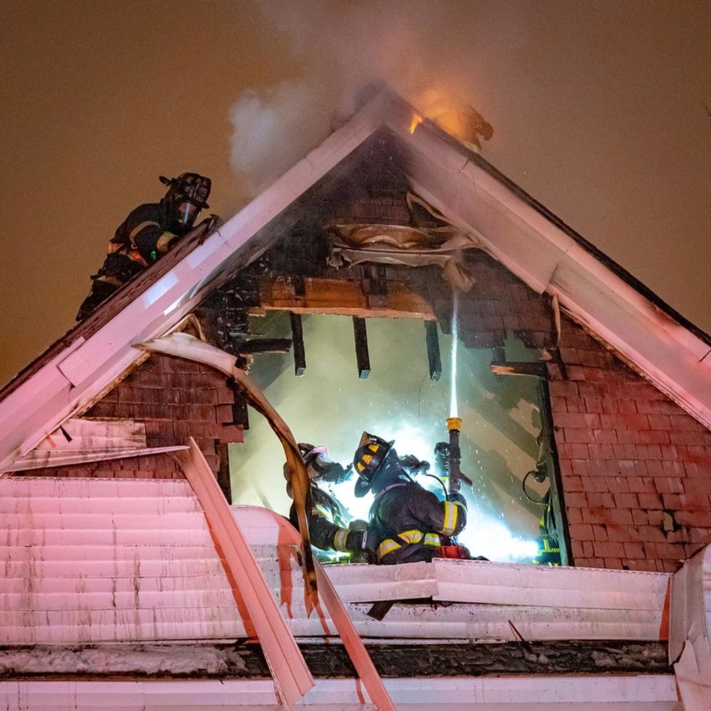 Firefighters, kneeling in an attic direct  a hose line toward the ceiling just under the roof.