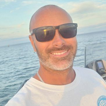 A smiling man wearing sunglasses on a boat with the ocean in the background.