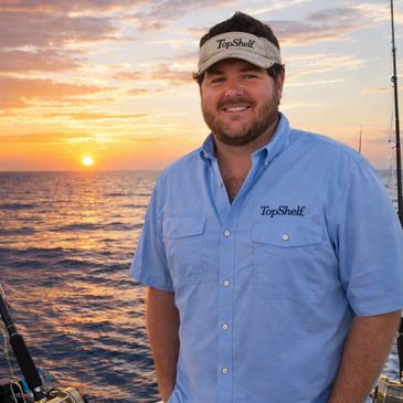 Man smiling on a boat during sunset, wearing a TopShelf shirt and visor.
