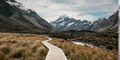 A winding wooden path leads through a grassy valley towards snow-capped mountains.