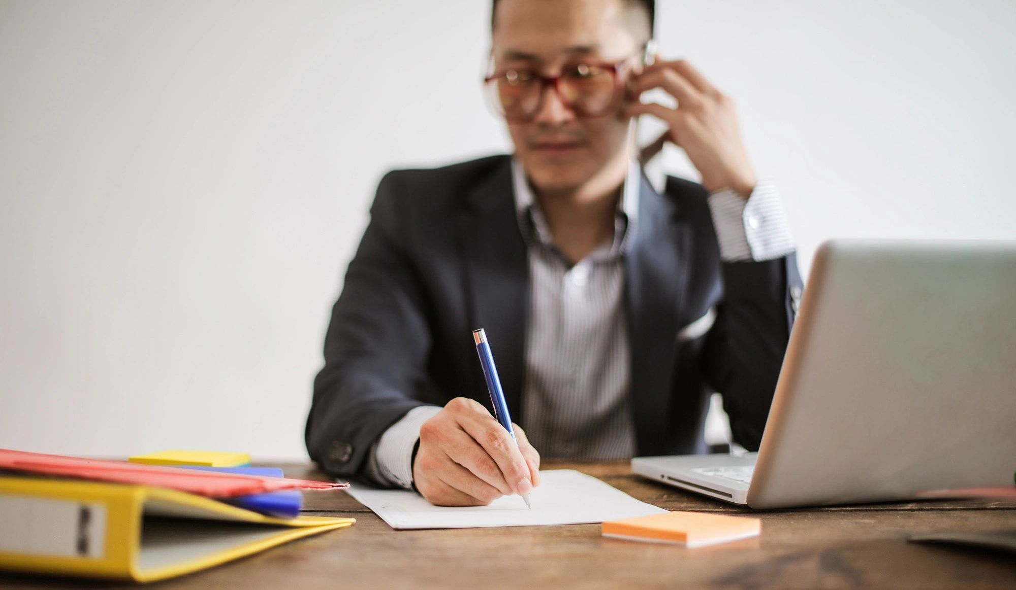 Businessman multitasking by writing and talking on phone at desk with laptop.