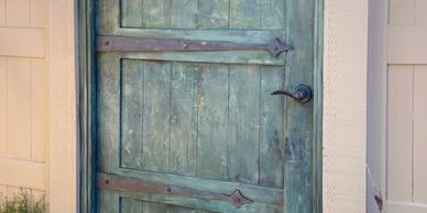 A weathered green wooden door with metal hinges on a beige wall.