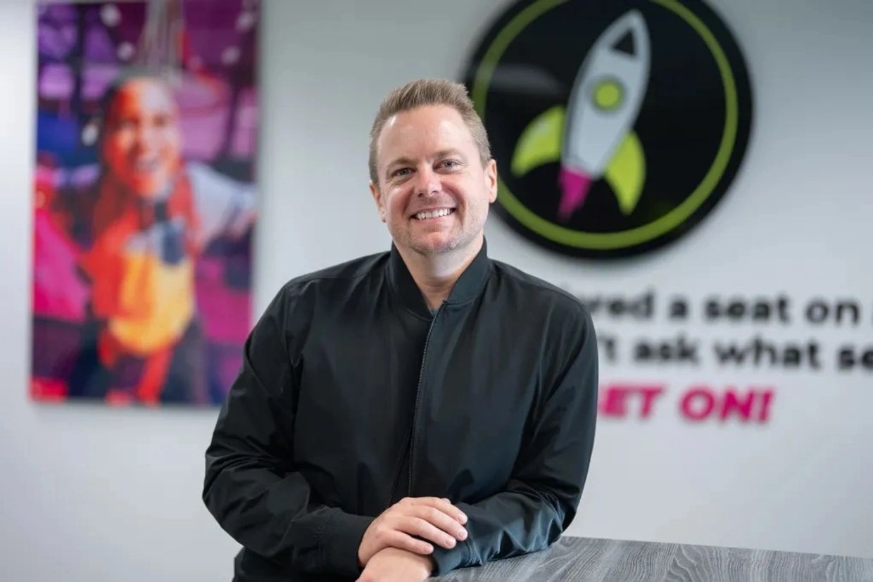 Man smiling in a black jacket with colorful blurred background.