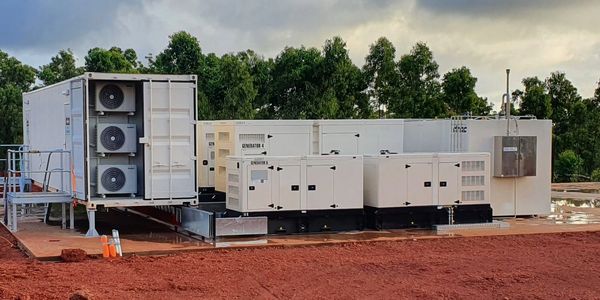 A set of large white industrial generators on a red dirt site with trees and cloudy sky.