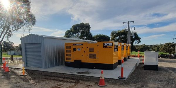 A row of yellow JCB generators beside a small shed on a sunny day.