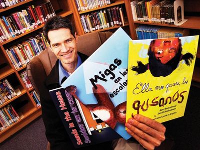 A man in a library holding three colorful books with Spanish titles.
