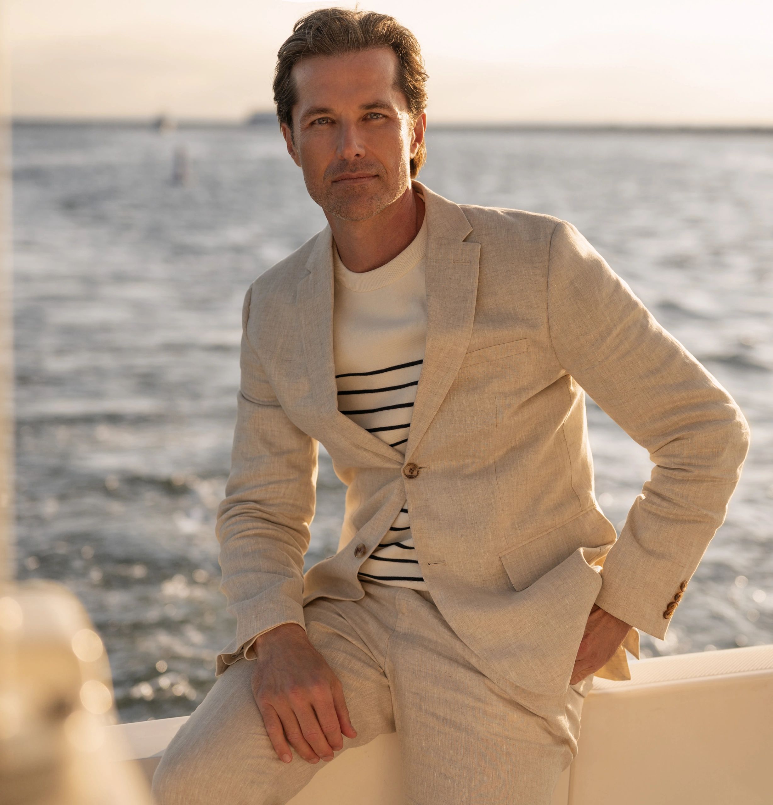 Man in beige suit by the water at sunset.