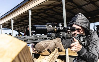 A man in a hoodie aiming a scoped rifle while resting on a wooden support.
