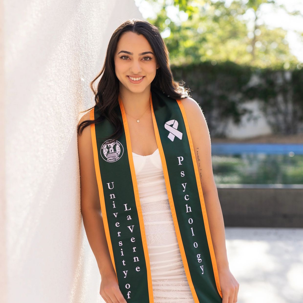 Young woman in graduation attire smiling outdoors against a white wall.