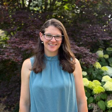 Smiling woman with glasses stands outdoors in front of greenery and flowers.
