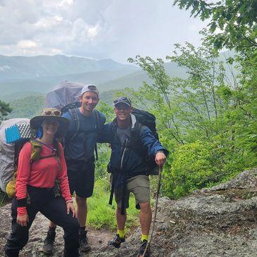 Three hikers posing on a mountain trail with lush greenery and misty mountains in the background.