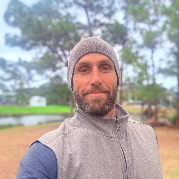Man in a gray beanie and vest smiling outdoors.