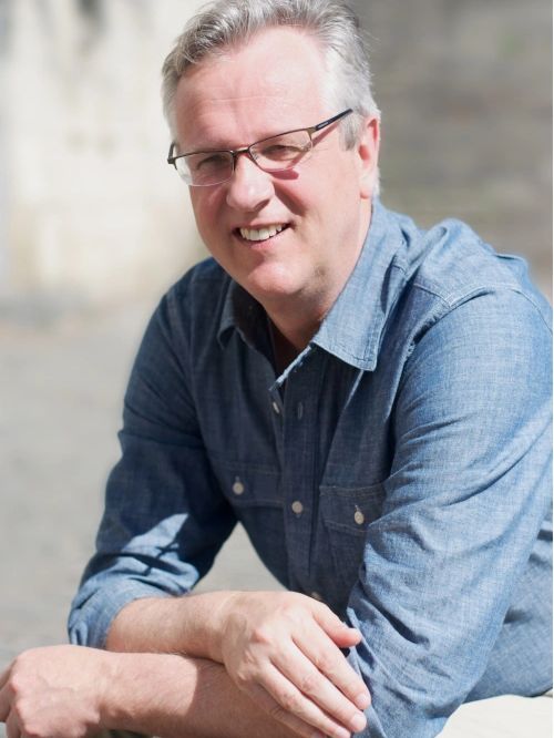 Smiling man wearing glasses and a blue shirt sitting outdoors.
