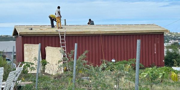 Three workers building a roof on a red container structure in a garden area.