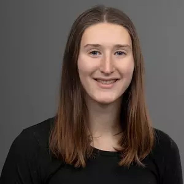 Young woman with straight brown hair smiling against a gray background.