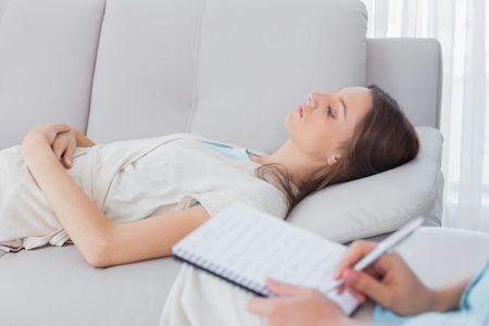 Woman hypnotherapy client reclining on a couch with therapist writing on a pad nearby.