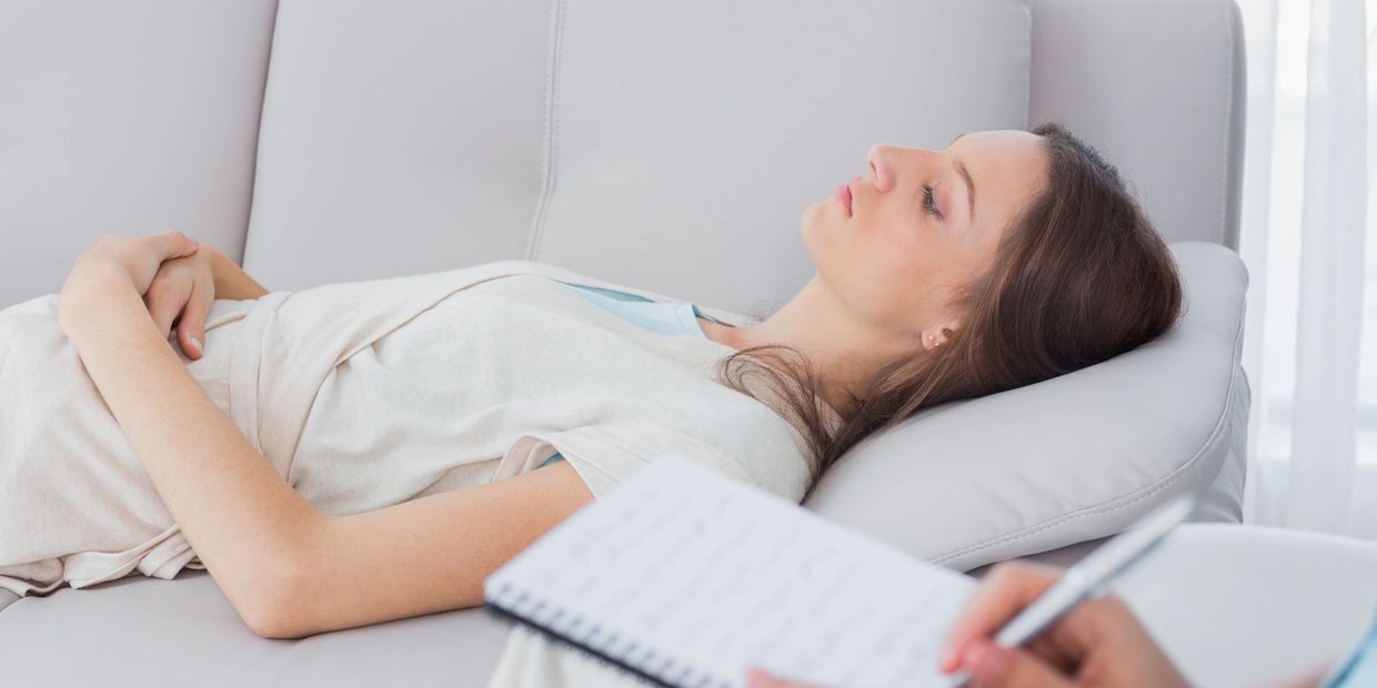 a woman lying on a couch with her eyes closed. someone sat next to them writing in a notepad