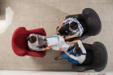 Three people in a meeting reviewing documents in chairs around a table.