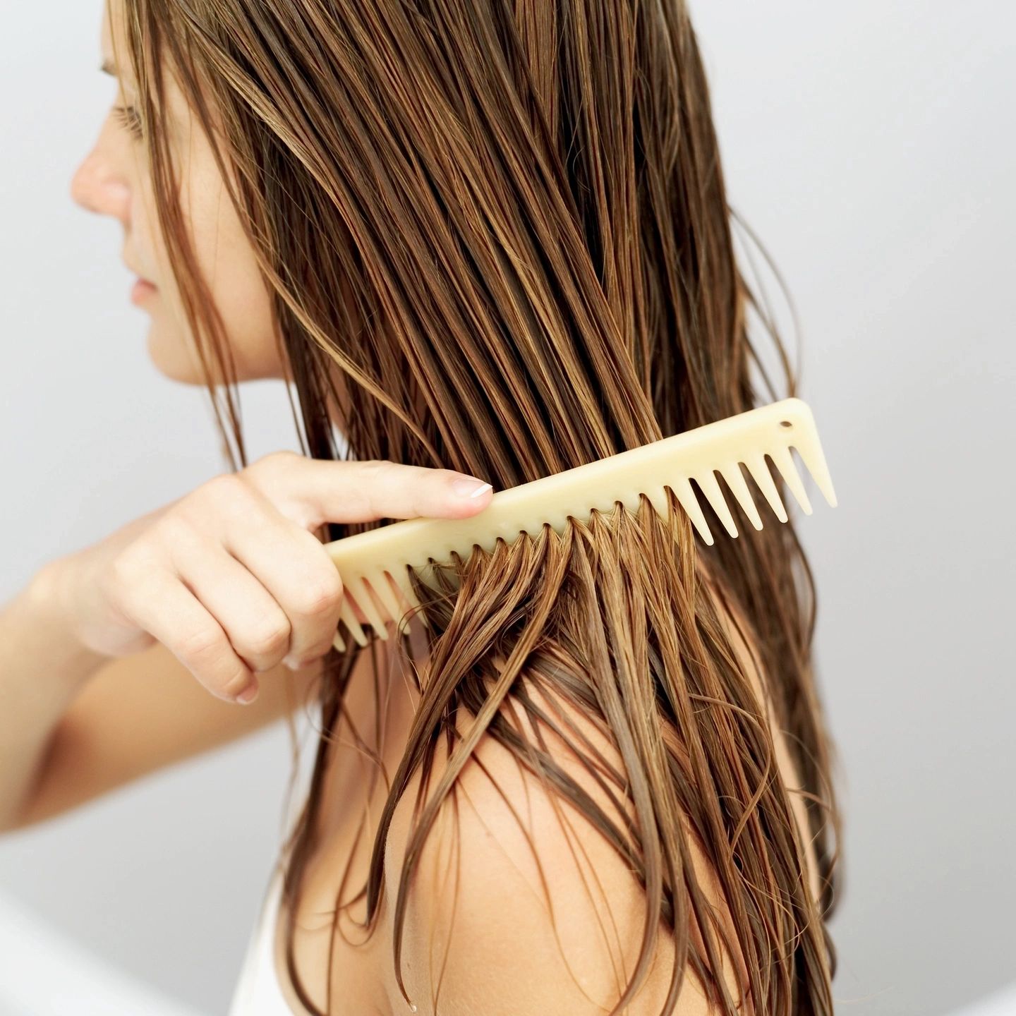 Side profile of a woman with brown hair using a tan-skin colored comb to brush her wet hair.