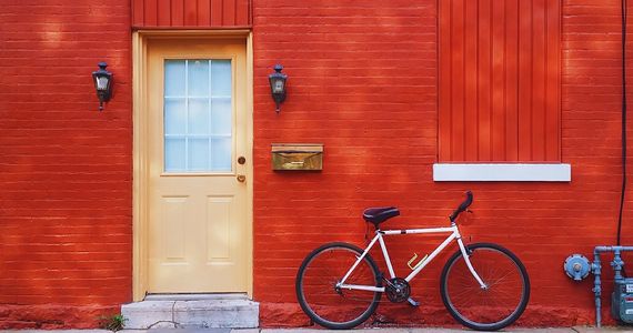 White bicycle leans against brick wall painted red, next to a pale yellow door.