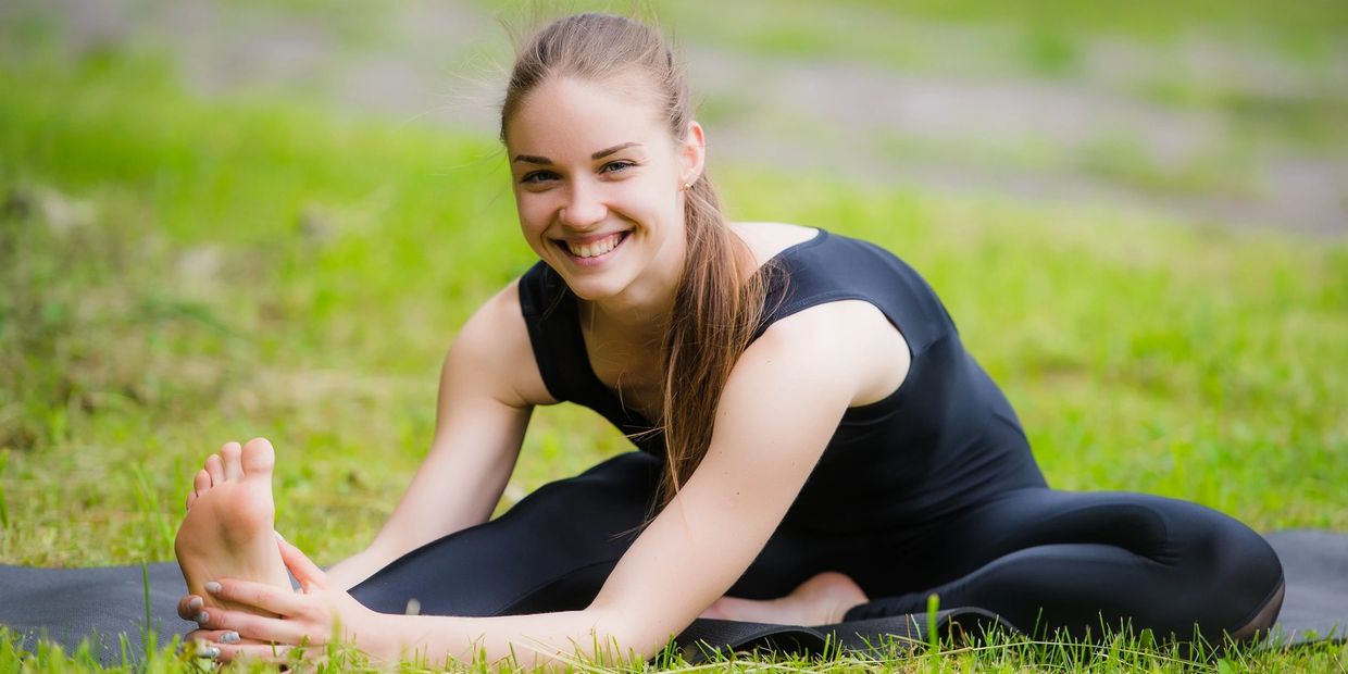 A smiling woman in black workout clothes stretches outdoors on a yoga mat.