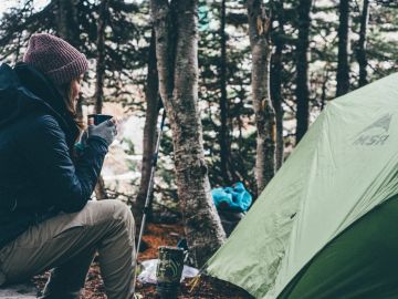 Person enjoying a hot drink near a green tent in the woods.