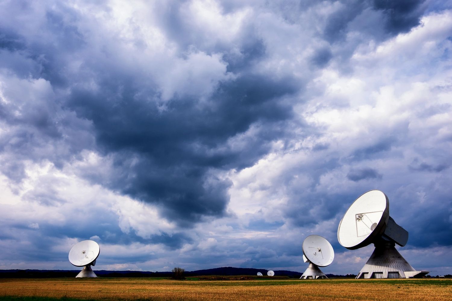 Satellite dishes under a dramatic cloudy sky in an open field.