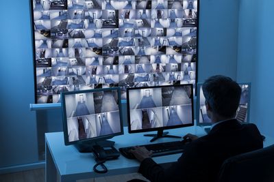A man at a computer monitoring many security camera feeds in a dimly lit room