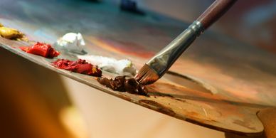 Close-up of a paintbrush mixing brown paint on a palette with various other colors.