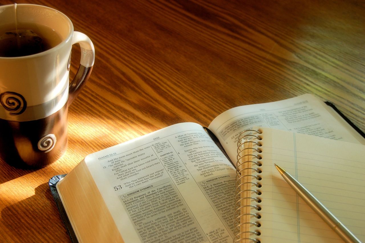 A tall cup of tea with a brew bag, a book and a notebook, sit on a table.