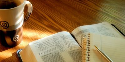 Open book, notebook, pen, and coffee cup on a wooden table.