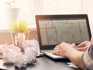 Person working on architectural floor plans on a laptop with crumpled papers nearby.