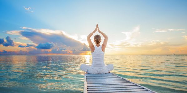 Person meditating at the end of an ocean dock with the sun setting in the distance.