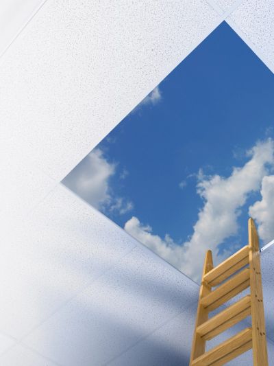 Wooden ladder reaching through ceiling tiles to a bright blue sky.