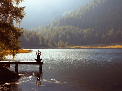 Person meditating on a dock by a serene lake surrounded by trees and mountains.
