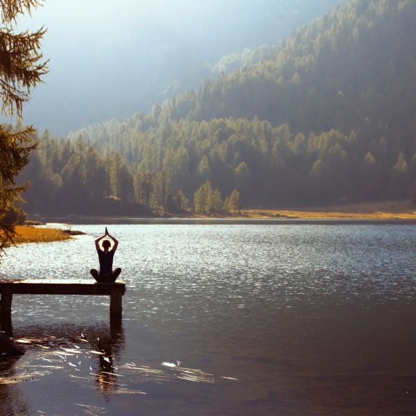 Person meditating on a dock by a serene lake with forested mountains.