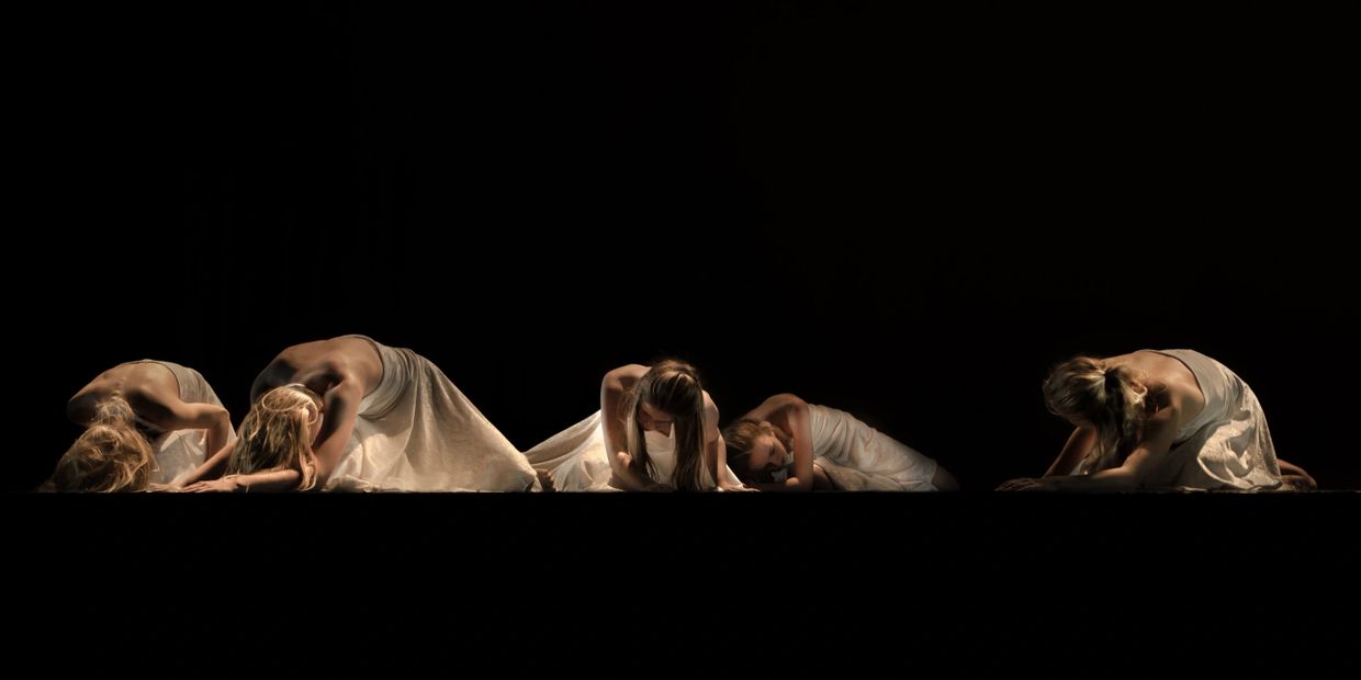 Five dancers in white dresses pose gracefully in a dimly lit performance.