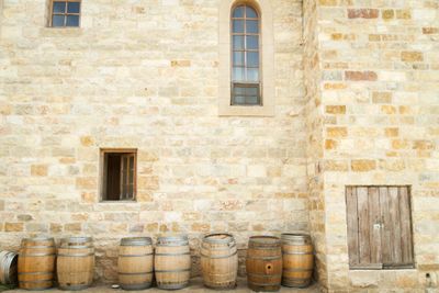 Wooden barrels lined up against a stone wall with small windows and a wooden door.