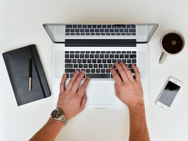 Hands typing on a laptop with coffee, phone, and notebook nearby.