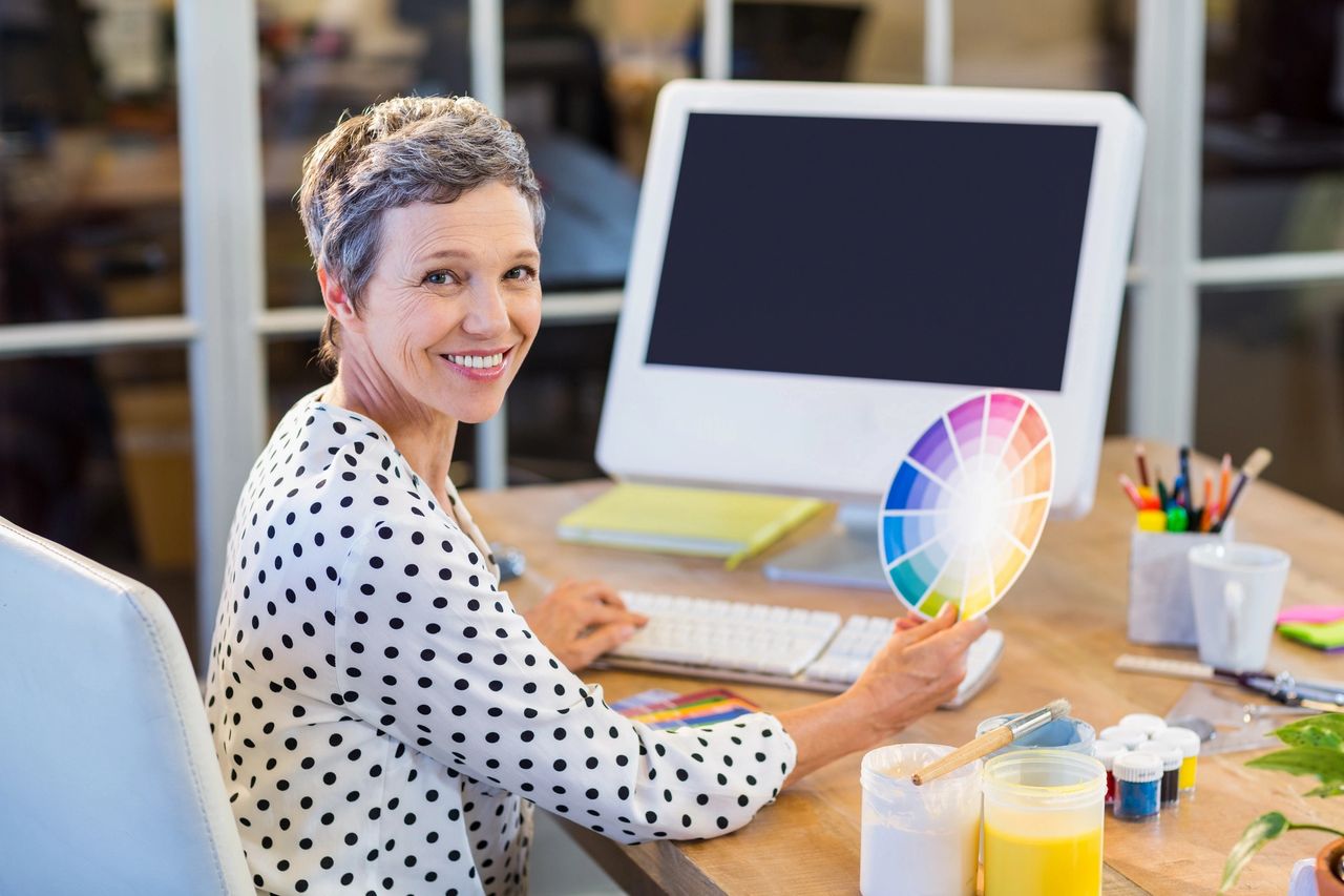 Woman smiles as she holds a diverse color wheel, with the paints on her desk and typing on her computer keyboard showing her expertise.