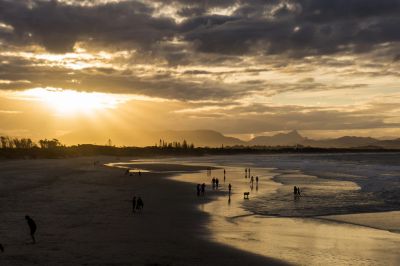 People walking along a beach at sunset with dramatic clouds and mountains in the distance.