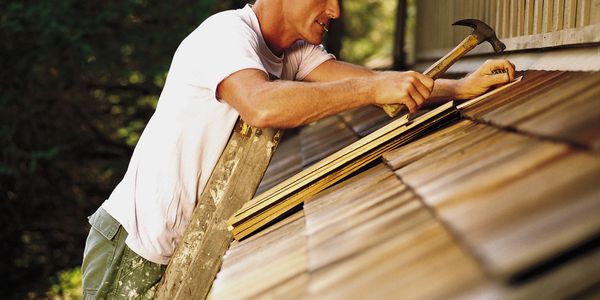 A man repairs a roof using a hammer while standing on a ladder.