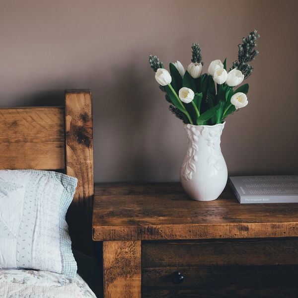 White flowers in a vase on a wooden bedside table next to a bed.