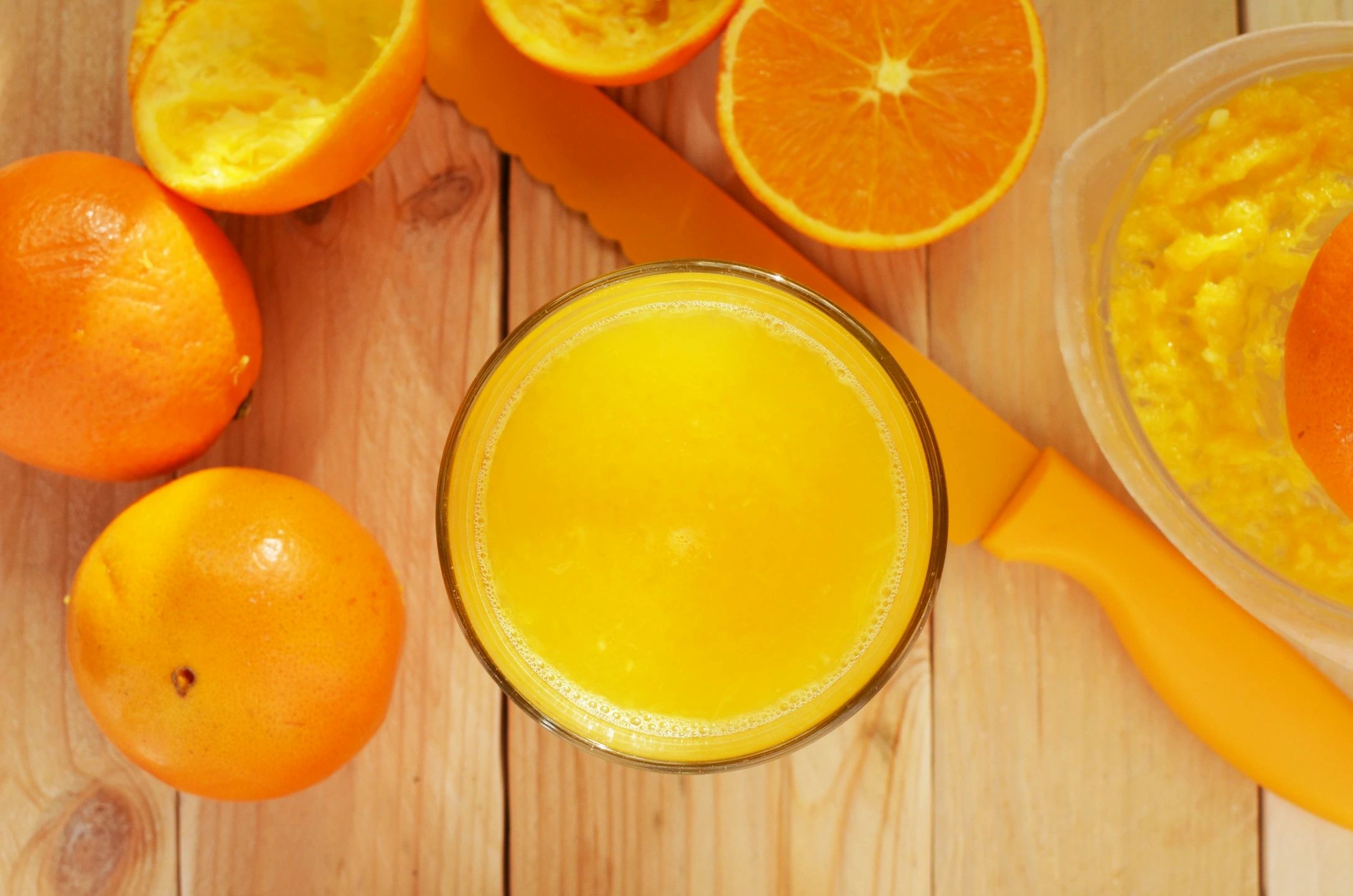 A glass of orange juice is on a wooden table next to oranges.